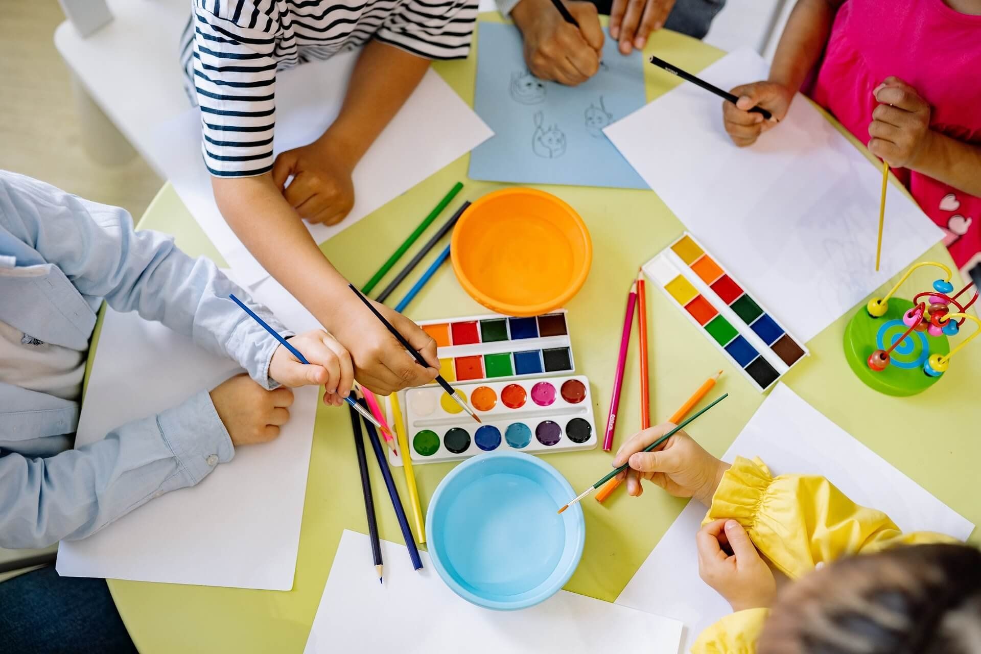 Children painting with watercolours at a yellow table covered in paint palettes, brushes and paper.