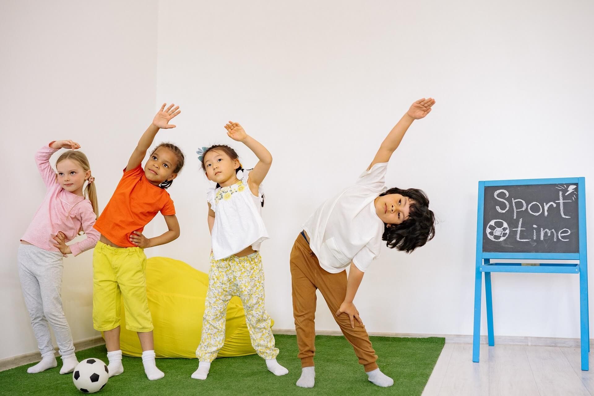 Four young children and a teacher stretching side to side in a bright playroom.
