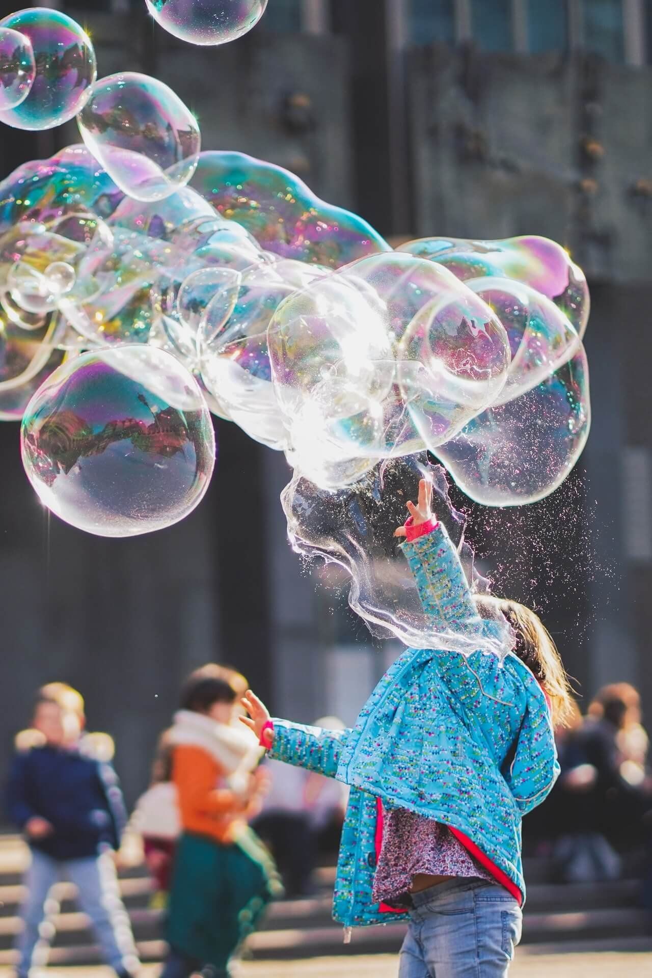 A young girl in a blue jacket blowing soap bubbles outdoors with other children behind her.