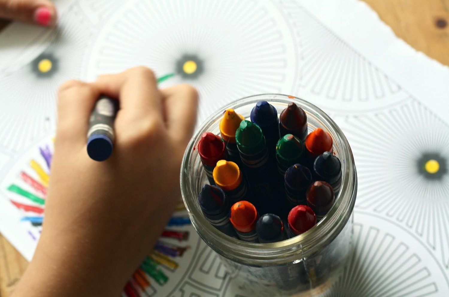 A child’s hand holding a blue crayon and colouring a sheet next to a jar of crayons.
