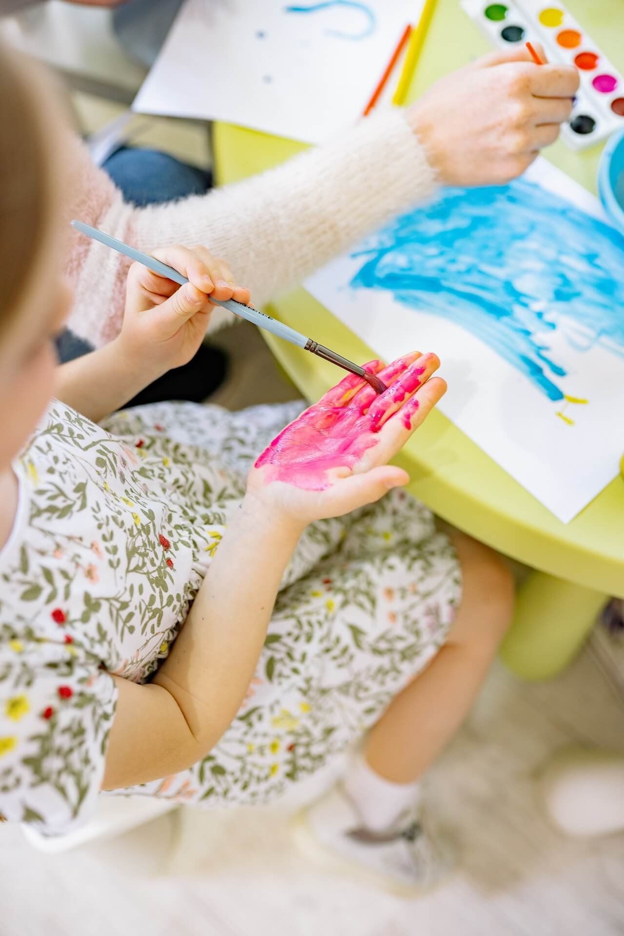 Child painting with pink and orange paint at a yellow table with watercolours and paper.