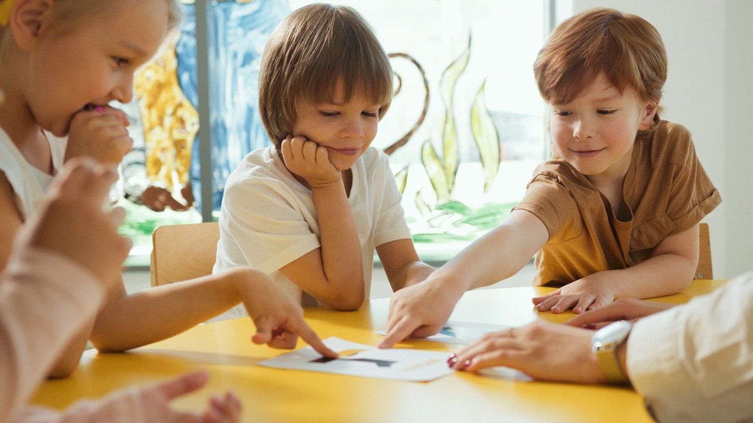 Children sitting around a yellow table pointing at a paper with the letter A while an adult helps.