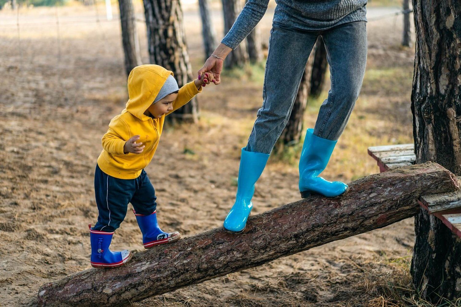 A young child in a yellow hoodie and rain boots holding an adult’s hand while balancing along a log in the woods.
