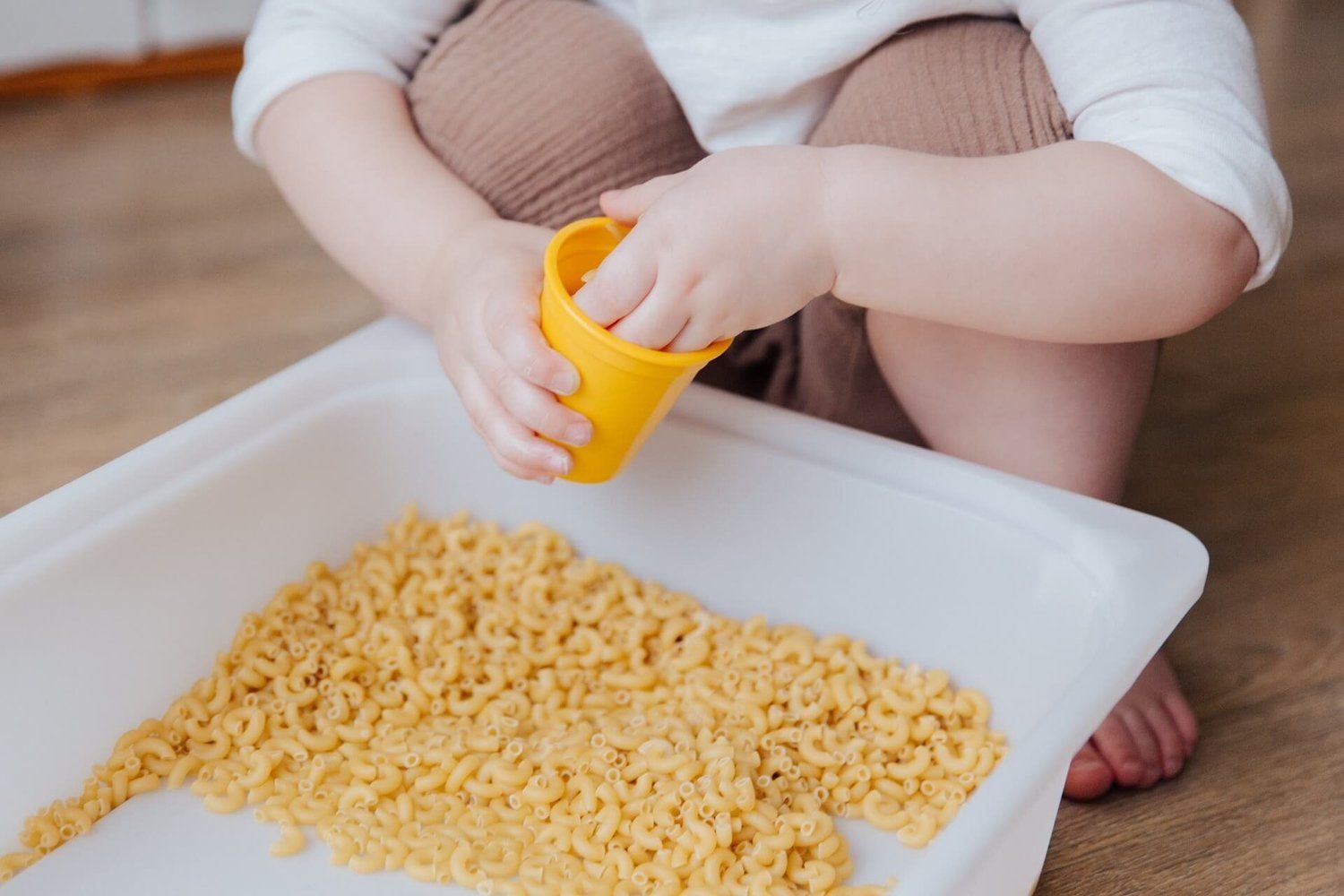 Child kneeling on a wooden floor playing with yellow play dough over a tray of uncooked pasta.