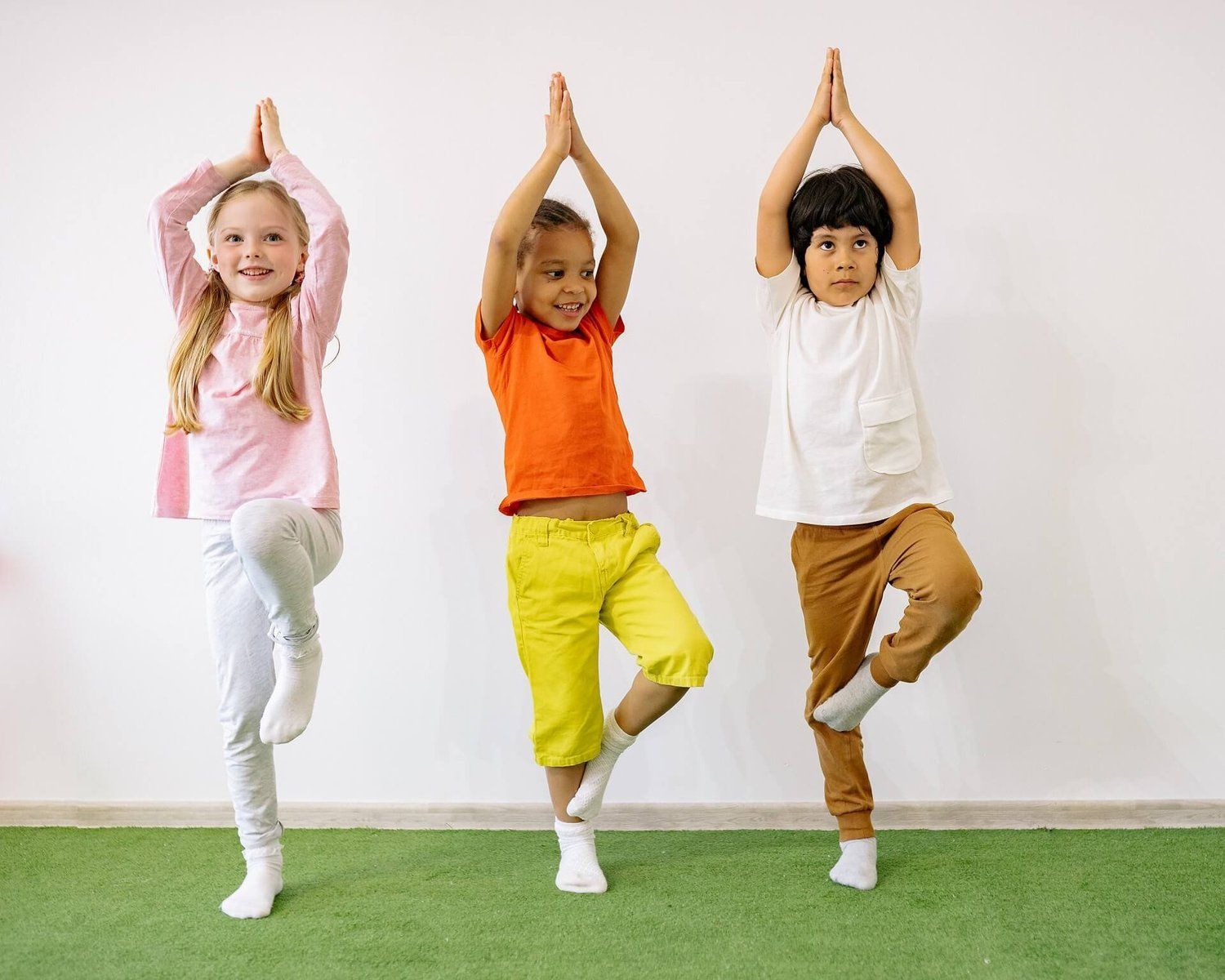 Three children practising yoga indoors, standing on one leg with hands together above their heads on a green mat.